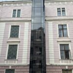 Зовнішній панорамний ліфт Izamet для памʼятки архітектури в Чернівцях — фото 1 Скляна шахта панорамного ліфта на темному металокаркасі фронтально на всю висоту дворового фасаду чотириповерхової будівлі